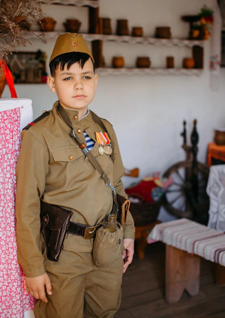 A child dressed in a vintage military uniform stands in a rustic room that appears to be historically themed. The room is decorated with traditional household items such as pottery, a samovar, and embroidered tablecloths. The setting evokes a sense of nostalgia and historical reenactment.