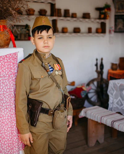 A child dressed in a vintage military uniform stands in a rustic room that appears to be historically themed. The room is decorated with traditional household items such as pottery, a samovar, and embroidered tablecloths. The setting evokes a sense of nostalgia and historical reenactment.