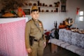 A child dressed in a vintage military uniform stands in a rustic room that appears to be historically themed. The room is decorated with traditional household items such as pottery, a samovar, and embroidered tablecloths. The setting evokes a sense of nostalgia and historical reenactment.