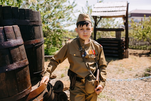 A child in a military uniform is standing outdoors beside a large wooden barrel. The uniform includes several medals, and the child is wearing a cap. In the background, there is a wooden structure and a fence, with green foliage suggesting it is spring or summer.