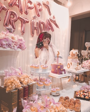 A festive birthday setting featuring a young woman joyfully holding a gift. The backdrop has large, pink balloon letters spelling 'HAPPY BIRTHDAY'. The celebration table is adorned with elegantly packaged treats and pastries, including cupcakes and a multi-tiered cake decorated with celestial motifs. Soft, pink floral arrangements complement the scene, and a warm, inviting glow emanates from surrounding lights.