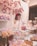 A festive birthday setting featuring a young woman joyfully holding a gift. The backdrop has large, pink balloon letters spelling 'HAPPY BIRTHDAY'. The celebration table is adorned with elegantly packaged treats and pastries, including cupcakes and a multi-tiered cake decorated with celestial motifs. Soft, pink floral arrangements complement the scene, and a warm, inviting glow emanates from surrounding lights.