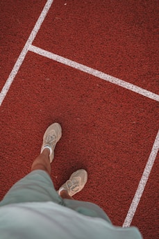 A person is standing on a red running track with white lane lines. The person is wearing light-colored sneakers and green shorts.