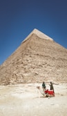 A large pyramid rises majestically under a clear blue sky. At the base, two people stand near a camel that is resting on the sandy ground. The pyramid is constructed of large stone blocks and creates a sense of ancient grandeur.