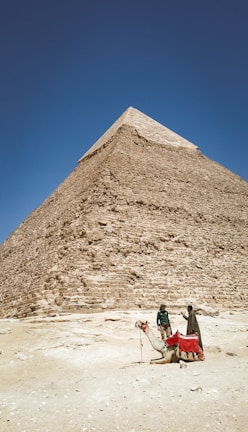A large pyramid rises majestically under a clear blue sky. At the base, two people stand near a camel that is resting on the sandy ground. The pyramid is constructed of large stone blocks and creates a sense of ancient grandeur.
