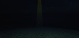 A dark field is illuminated by a single beam of yellowish light in the center. The light creates a mysterious or eerie atmosphere, highlighting the grass in front while the background remains obscured.