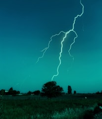 A bright lightning bolt strikes down from the sky over a flat rural landscape. The scene is dominated by an expansive field and a few scattered trees and bushes in the background. The sky has a deep blue-green hue, giving the scene an otherworldly appearance.