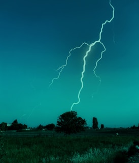 A bright lightning bolt strikes down from the sky over a flat rural landscape. The scene is dominated by an expansive field and a few scattered trees and bushes in the background. The sky has a deep blue-green hue, giving the scene an otherworldly appearance.