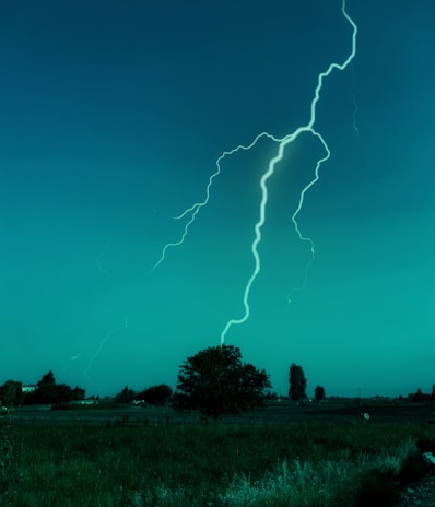 A bright lightning bolt strikes down from the sky over a flat rural landscape. The scene is dominated by an expansive field and a few scattered trees and bushes in the background. The sky has a deep blue-green hue, giving the scene an otherworldly appearance.