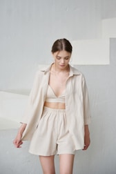 A young woman with brown hair is wearing a light beige outfit consisting of a loose shirt over a top and shorts, standing against a minimalist, pale background with stairs. Her hair is tied back, and she has hoop earrings and a cross necklace.