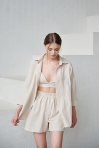 A young woman with brown hair is wearing a light beige outfit consisting of a loose shirt over a top and shorts, standing against a minimalist, pale background with stairs. Her hair is tied back, and she has hoop earrings and a cross necklace.