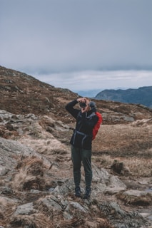 A person is standing on a rocky, grassy hilltop with a camera, set against a backdrop of overcast skies and distant mountains. The individual is dressed in outdoor clothing and a red backpack, suggesting a hiking excursion.