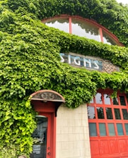 A building facade covered in lush green ivy with a red door and window frames. Above the door, a wooden sign displays the word 'SIGNS' in a stylized font. The structure has a vintage and natural aesthetic, highlighted by the combination of vibrant greenery and warm red tones.