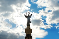 A silhouetted statue of an angel holding two wreaths atop a tall pedestal, set against a partly cloudy sky with the sun peeking through the clouds.