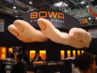 A trade show booth featuring a large installation resembling a pair of oversized hands positioned above. The booth is branded with the name 'BOWA' and several display cases with products are illuminated in the background. Several individuals are present, engaging in conversation and looking at the exhibits.