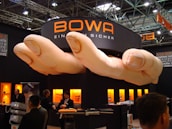 A trade show booth featuring a large installation resembling a pair of oversized hands positioned above. The booth is branded with the name 'BOWA' and several display cases with products are illuminated in the background. Several individuals are present, engaging in conversation and looking at the exhibits.