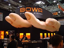 A trade show booth featuring a large installation resembling a pair of oversized hands positioned above. The booth is branded with the name 'BOWA' and several display cases with products are illuminated in the background. Several individuals are present, engaging in conversation and looking at the exhibits.