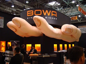 A trade show booth featuring a large installation resembling a pair of oversized hands positioned above. The booth is branded with the name 'BOWA' and several display cases with products are illuminated in the background. Several individuals are present, engaging in conversation and looking at the exhibits.