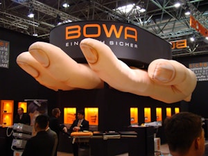 A trade show booth featuring a large installation resembling a pair of oversized hands positioned above. The booth is branded with the name 'BOWA' and several display cases with products are illuminated in the background. Several individuals are present, engaging in conversation and looking at the exhibits.