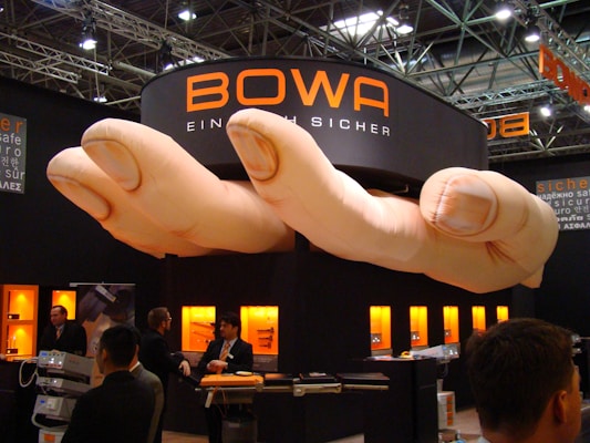 A trade show booth featuring a large installation resembling a pair of oversized hands positioned above. The booth is branded with the name 'BOWA' and several display cases with products are illuminated in the background. Several individuals are present, engaging in conversation and looking at the exhibits.