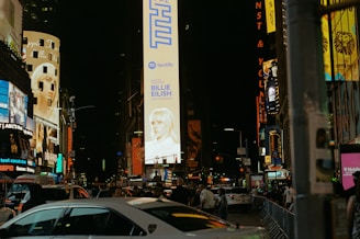 Brightly lit urban scene showcasing towering billboards and bustling crowds on a night street. Large electronic advertisements feature prominently along the buildings, with cars and people moving along the road and sidewalks.