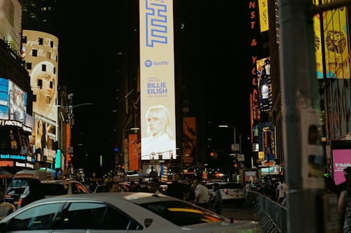 Brightly lit urban scene showcasing towering billboards and bustling crowds on a night street. Large electronic advertisements feature prominently along the buildings, with cars and people moving along the road and sidewalks.