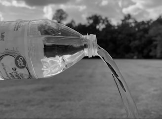 A plastic water bottle is tilted downward, pouring out water against a blurred outdoor background featuring trees and a cloudy sky. The scene is in black and white.
