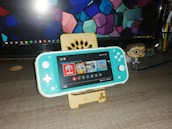 A turquoise handheld gaming console is placed on a wooden stand in front of a computer monitor displaying geometric shapes. To the right, there is a small figurine of a person wearing glasses and a suit. The gaming console screen shows a menu with various game icons.