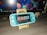 A turquoise handheld gaming console is placed on a wooden stand in front of a computer monitor displaying geometric shapes. To the right, there is a small figurine of a person wearing glasses and a suit. The gaming console screen shows a menu with various game icons.