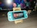 A turquoise handheld gaming console is placed on a wooden stand in front of a computer monitor displaying geometric shapes. To the right, there is a small figurine of a person wearing glasses and a suit. The gaming console screen shows a menu with various game icons.