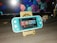 A turquoise handheld gaming console is placed on a wooden stand in front of a computer monitor displaying geometric shapes. To the right, there is a small figurine of a person wearing glasses and a suit. The gaming console screen shows a menu with various game icons.