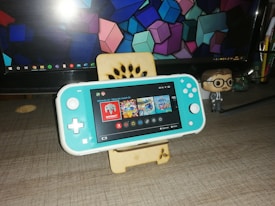 A turquoise handheld gaming console is placed on a wooden stand in front of a computer monitor displaying geometric shapes. To the right, there is a small figurine of a person wearing glasses and a suit. The gaming console screen shows a menu with various game icons.