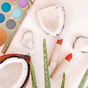 Slices of coconut and aloe leaves are arranged alongside a makeup palette featuring vibrant colors, including blue and purple shades. Two lipsticks in shades of red and nude are placed next to each other.