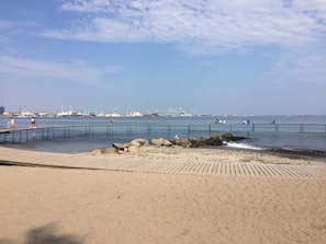 A sandy beach with a wooden boardwalk leading to a metal walkway that stretches over the calm sea. Several people are walking or sitting on the walkway, and a few seagulls are visible. The background features an industrial skyline with cranes and buildings under a blue sky with scattered clouds.
