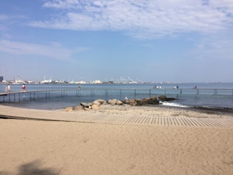 A sandy beach with a wooden boardwalk leading to a metal walkway that stretches over the calm sea. Several people are walking or sitting on the walkway, and a few seagulls are visible. The background features an industrial skyline with cranes and buildings under a blue sky with scattered clouds.