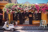 A charming storefront with a vibrant display of flowers cascading above. The building exterior is painted in a warm yellow hue, with dark shutters flanking the entrance. A white scooter is parked in front, and a chalkboard sign encouraging shopping stands nearby. Inside, the shop is filled with artisanal products and decorative items.