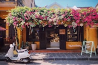 A charming storefront with a vibrant display of flowers cascading above. The building exterior is painted in a warm yellow hue, with dark shutters flanking the entrance. A white scooter is parked in front, and a chalkboard sign encouraging shopping stands nearby. Inside, the shop is filled with artisanal products and decorative items.