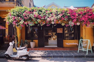 A charming storefront with a vibrant display of flowers cascading above. The building exterior is painted in a warm yellow hue, with dark shutters flanking the entrance. A white scooter is parked in front, and a chalkboard sign encouraging shopping stands nearby. Inside, the shop is filled with artisanal products and decorative items.