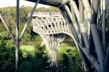A modern architectural structure made of interwoven metal beams that span across a lush green landscape. The structure includes a pedestrian walkway on top, with people visible standing and walking. The background features dense trees and greenery.