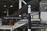 A black directional signpost with multiple signs pointing in different directions. Each sign bears historical inscriptions related to horse racing events and figures, such as 'Piggott beats Francome 1985' and 'The Queen Mother visited Warwick 1983'. The background features a blurred view of a building and spectators, indicating a sports venue setting.