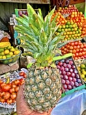 A hand holding a ripe pineapple in front of a fruit stall. The background features colorful stacks of various fruits including oranges, apples, and plums. The scene is vibrant and bustling with fresh produce.