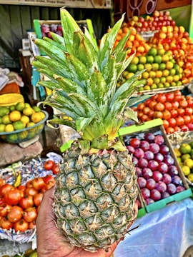 A hand holding a ripe pineapple in front of a fruit stall. The background features colorful stacks of various fruits including oranges, apples, and plums. The scene is vibrant and bustling with fresh produce.