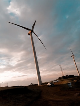 A landscape featuring several large wind turbines against a cloudy sky. The turbines are situated on a dirt terrain with vehicles parked nearby. The sun casts a warm, golden light, creating a serene and calm atmosphere.