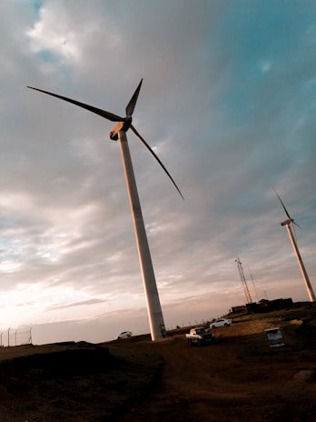 A landscape featuring several large wind turbines against a cloudy sky. The turbines are situated on a dirt terrain with vehicles parked nearby. The sun casts a warm, golden light, creating a serene and calm atmosphere.