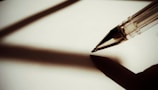 A ballpoint pen casts a deep shadow on a blank white surface. The pen is positioned at an angle, suggesting the potential to write.