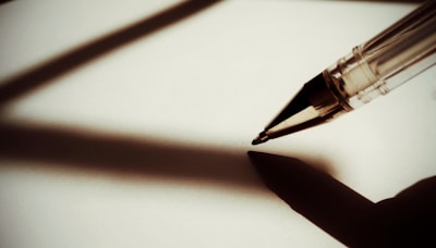 A ballpoint pen casts a deep shadow on a blank white surface. The pen is positioned at an angle, suggesting the potential to write.
