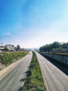 A wide, open highway with two lanes on each side bordered by greenery. On the left, there are buildings and a few vehicles, while the right side has a retaining wall with vegetation. The road appears relatively empty, with just a few cars driving under a clear blue sky.