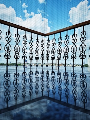 Ornate wrought iron railing with intricate patterns casts a reflection on a glossy surface, while a backdrop of blue sky with scattered clouds enhances the visual contrast.