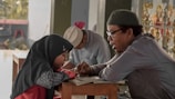 A man and a young girl engage in a focused discussion or study session. The girl, wearing a dark hijab, looks attentive as the man explains something to her. They are sitting at a wooden table, and the background shows another person in a light hat reading and some trophies displayed.