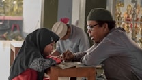 A man and a young girl engage in a focused discussion or study session. The girl, wearing a dark hijab, looks attentive as the man explains something to her. They are sitting at a wooden table, and the background shows another person in a light hat reading and some trophies displayed.
